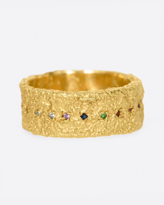 A yellow gold wide textured band set with garnet, ruby, peridot, aquamarine, amethyst, emerald, and pink, orange, yellow and blue sapphires. Shown from the front.