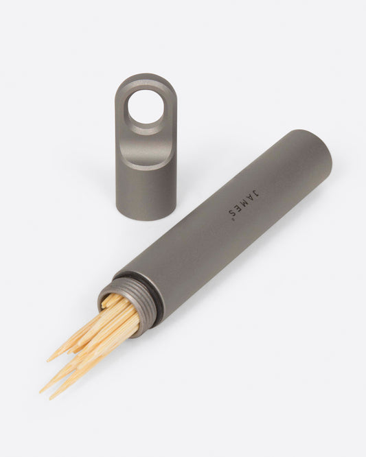 A narrow titanium stash canister shown lying on its side open with toothpicks inside. The lid is shown sitting up beside it.