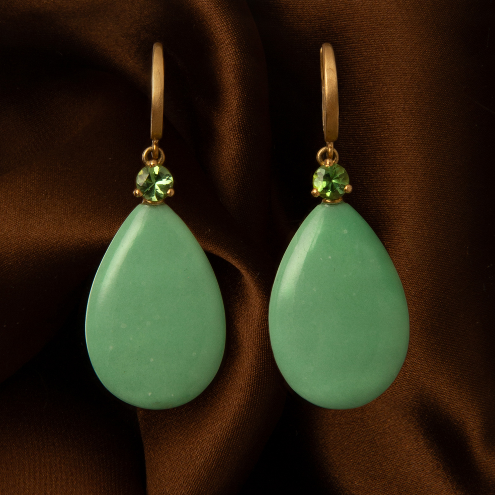 A pair of gold drop earrings with smooth, teardrop-shaped mint green stone pendants and small round green crystal accents, displayed against a draped dark brown satin fabric background.