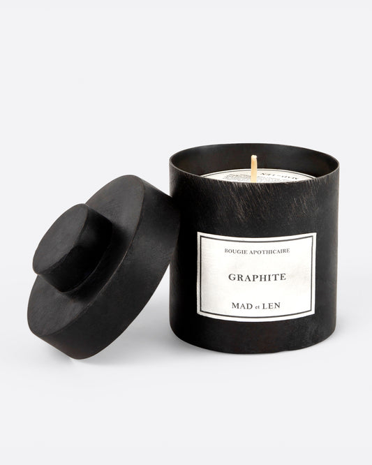 A Mad et Len bois Graphite candle measuring 3" tall and 3.25" wide. It is housed in a one-of-a-kind blackened iron vessel with an open lid and a label that reads: Bougie Apothicaire Graphite Mad et Len. Shown from the front.