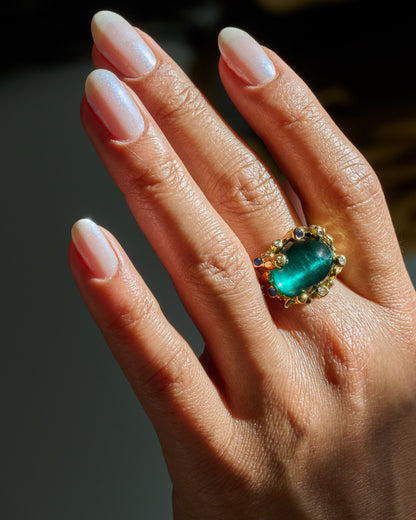 A yellow gold ring with an indicolite tourmaline cabochon encircled by blue sapphires and diamonds, shown on a hand. 