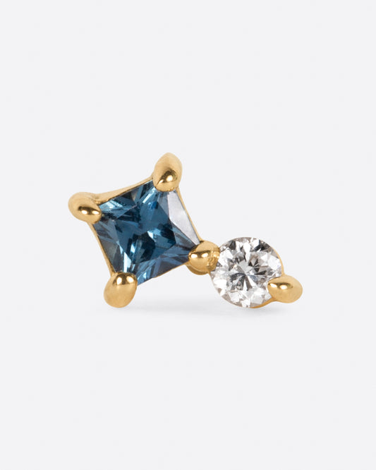 A yellow gold stud earring with a prong set round diamond sitting atop a princess cut teal blue sapphire. Shown on its side from the front.