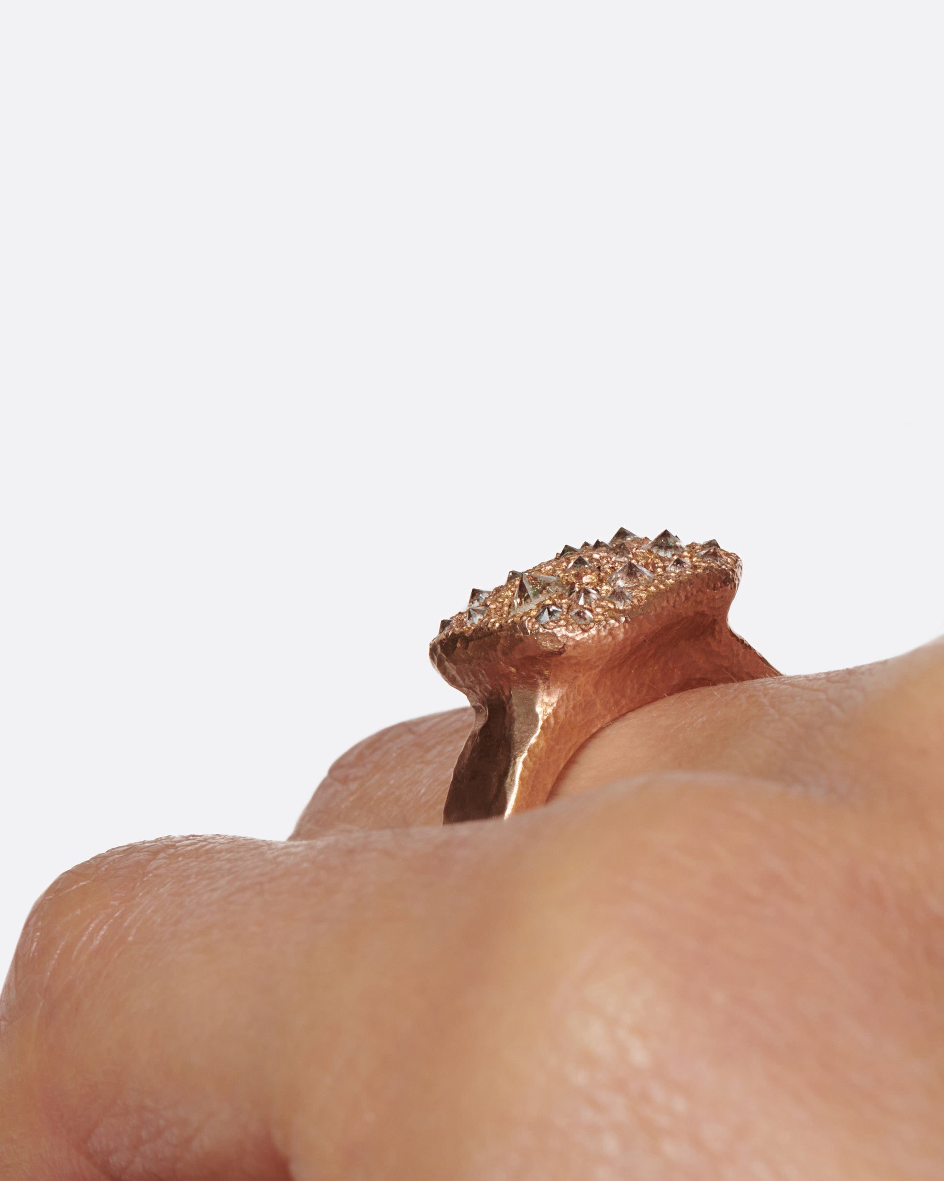 A hammered, hefty rose gold ring with inverted diamonds scattered across its rectangular face. Shown on a hand at an angle.