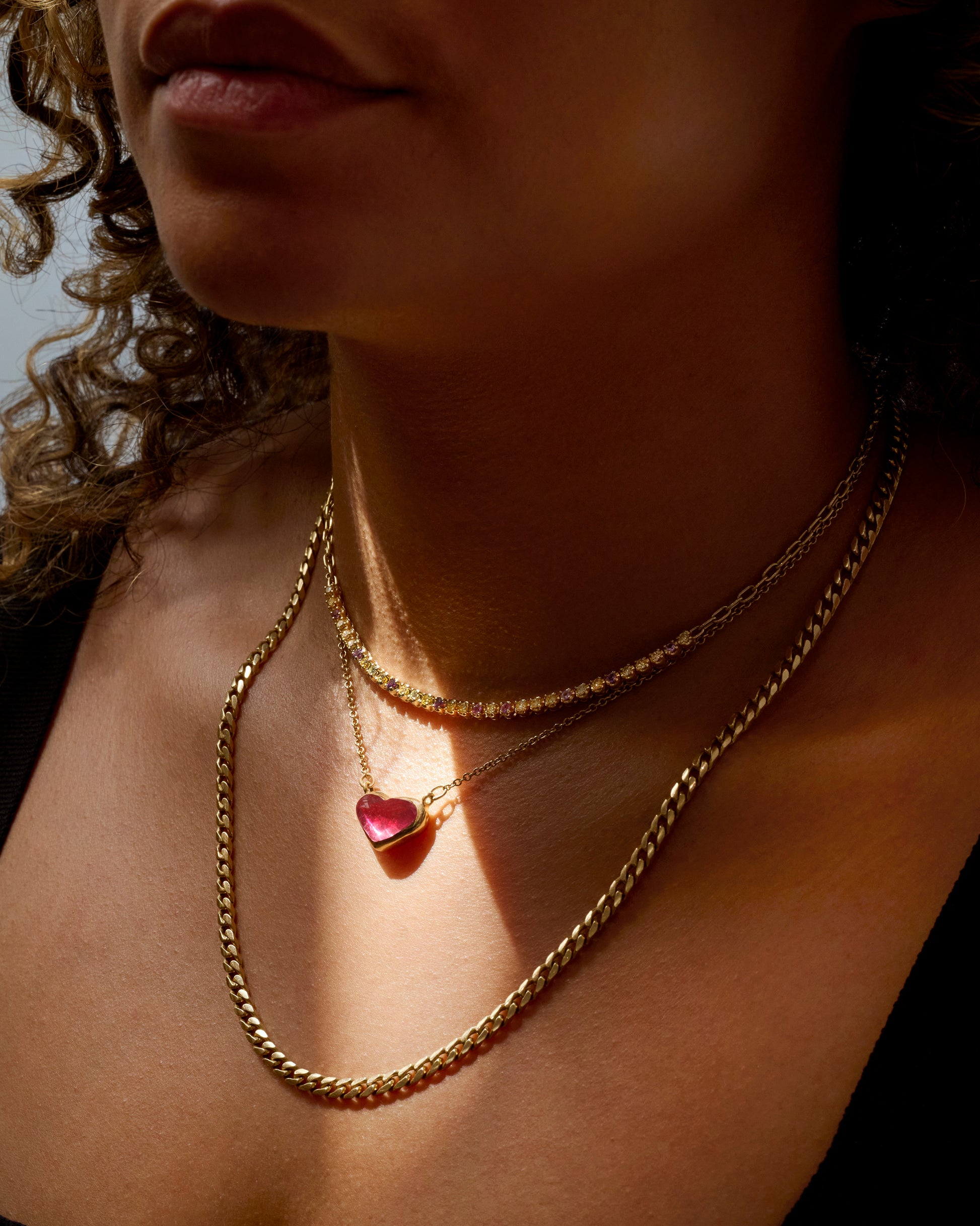 Three yellow gold necklaces- one with diamonds, one with a rubellite heart shaped pendant, and one thick chain, all shown on a person.