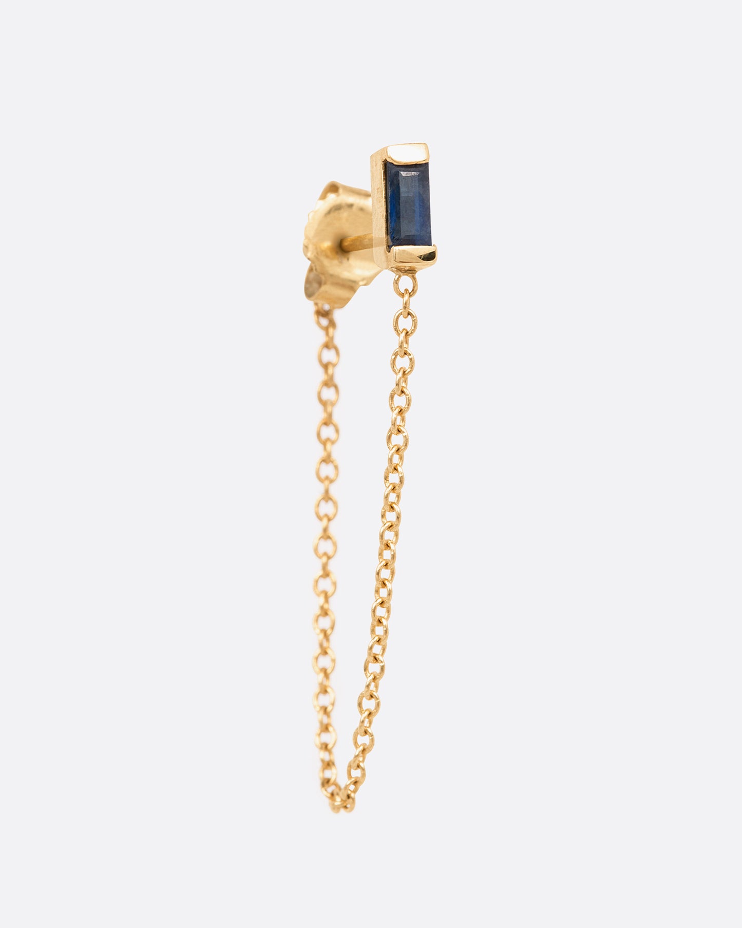 A yellow gold stud earring with a channel set sapphire baguette and a 1" chain drop looped. Shown from the front.