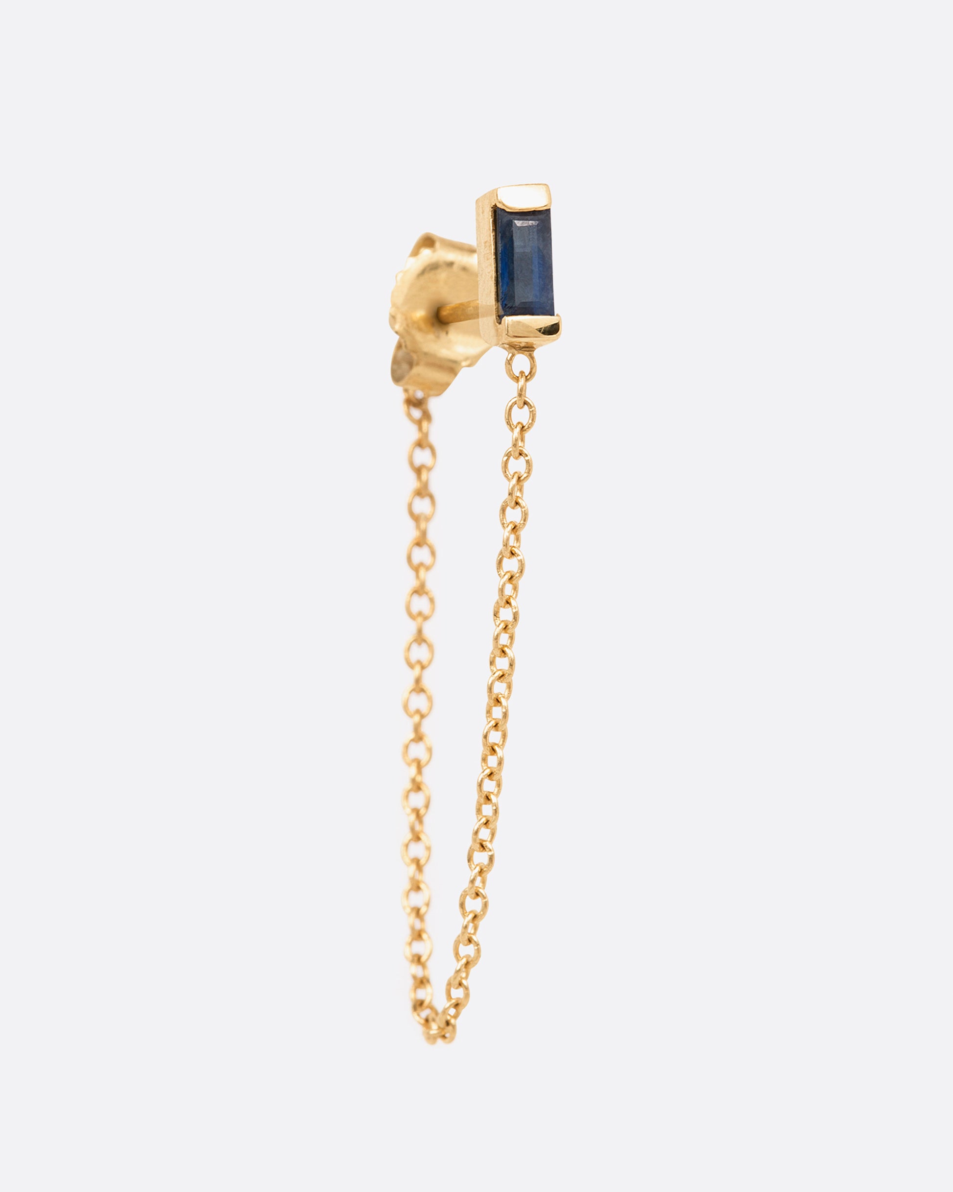 A yellow gold stud earring with a channel set sapphire baguette and a 1" chain drop looped. Shown from the front.