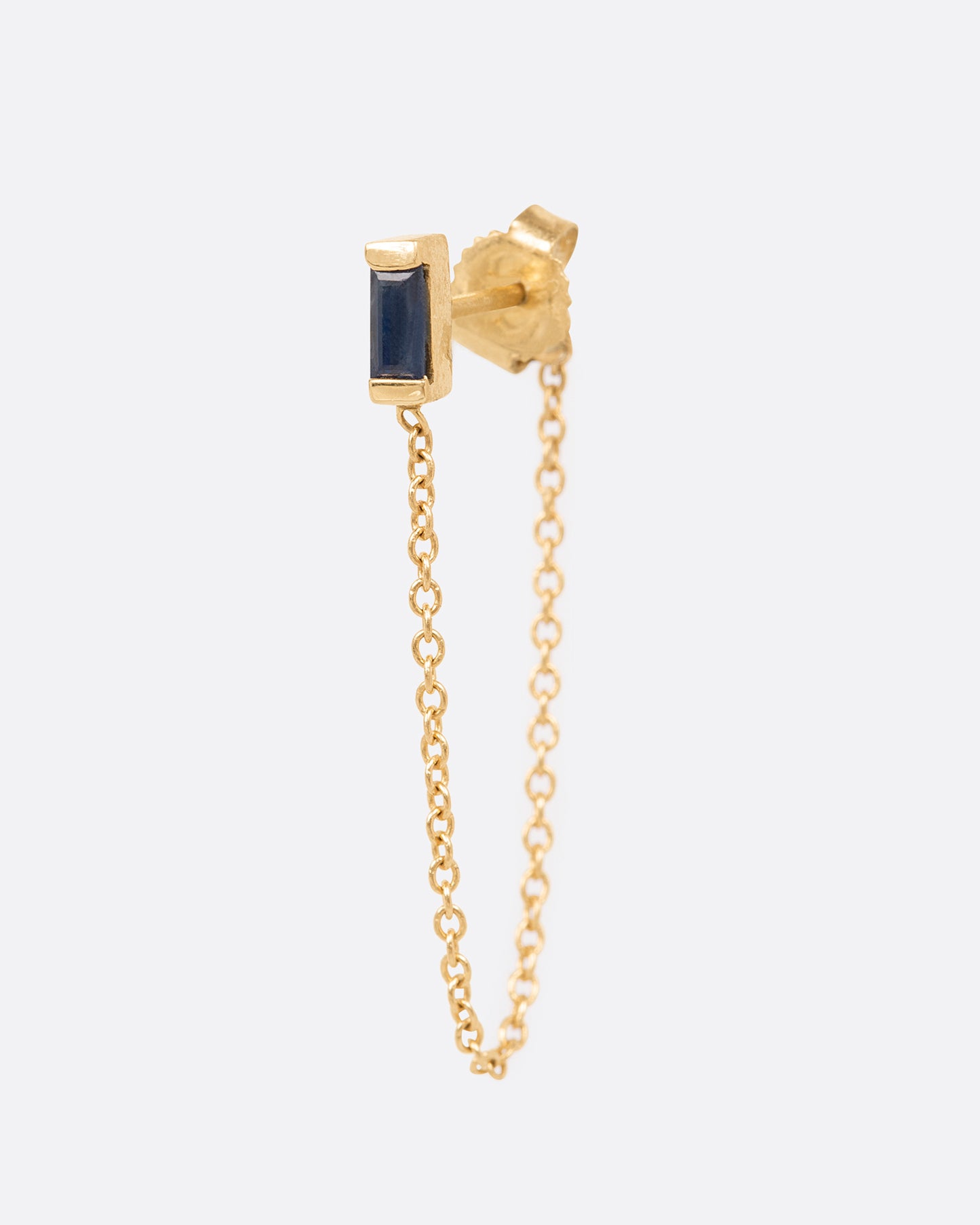 A yellow gold stud earring with a channel set sapphire baguette and a 1" chain drop looped. Shown from the side.