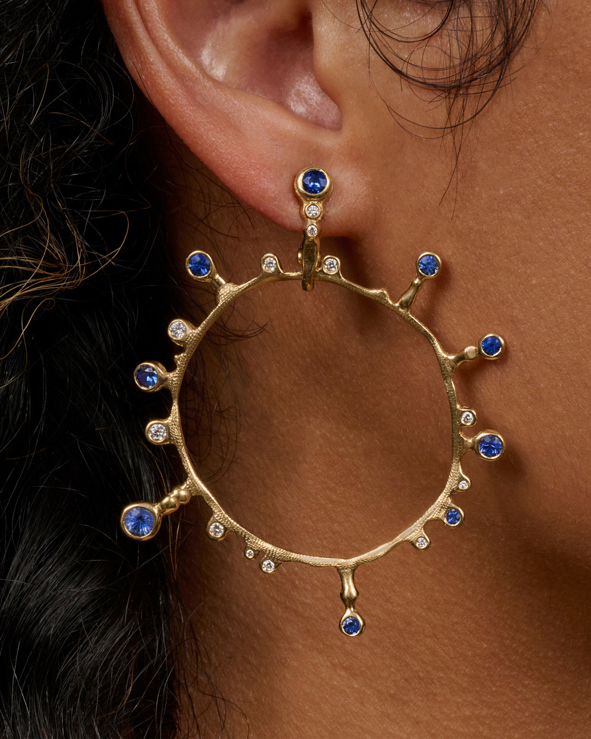 A yellow gold hoop sunburst earring with diamonds and sapphires. Shown from the front on a person.