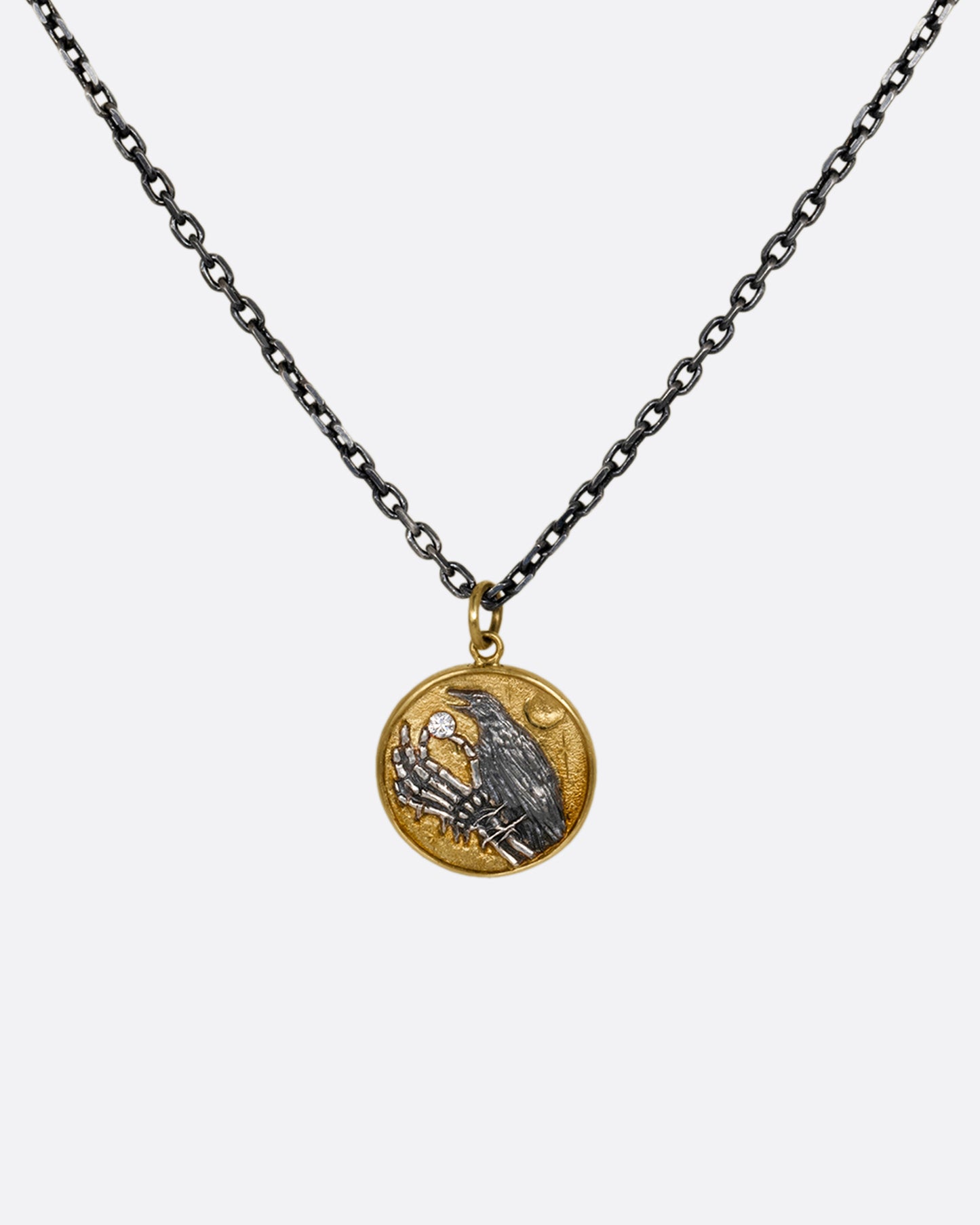 A yellow gold and silver pendant necklace with a skeleton hand holding a diamond and a crow. Shown from the front.