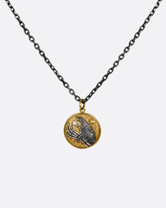 A yellow gold and silver pendant necklace with a skeleton hand holding a diamond and a crow. Shown from the front.