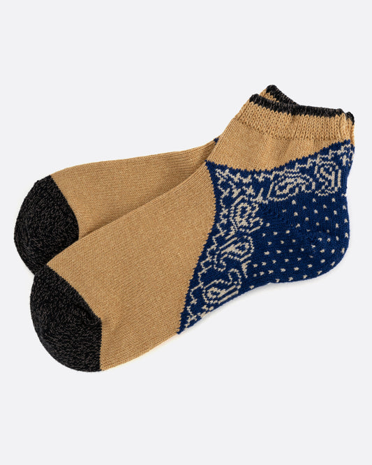 A pair of ochre and charcoal ankle socks with a navy blue paisley bandana print heel. Shown lying flat from the side.