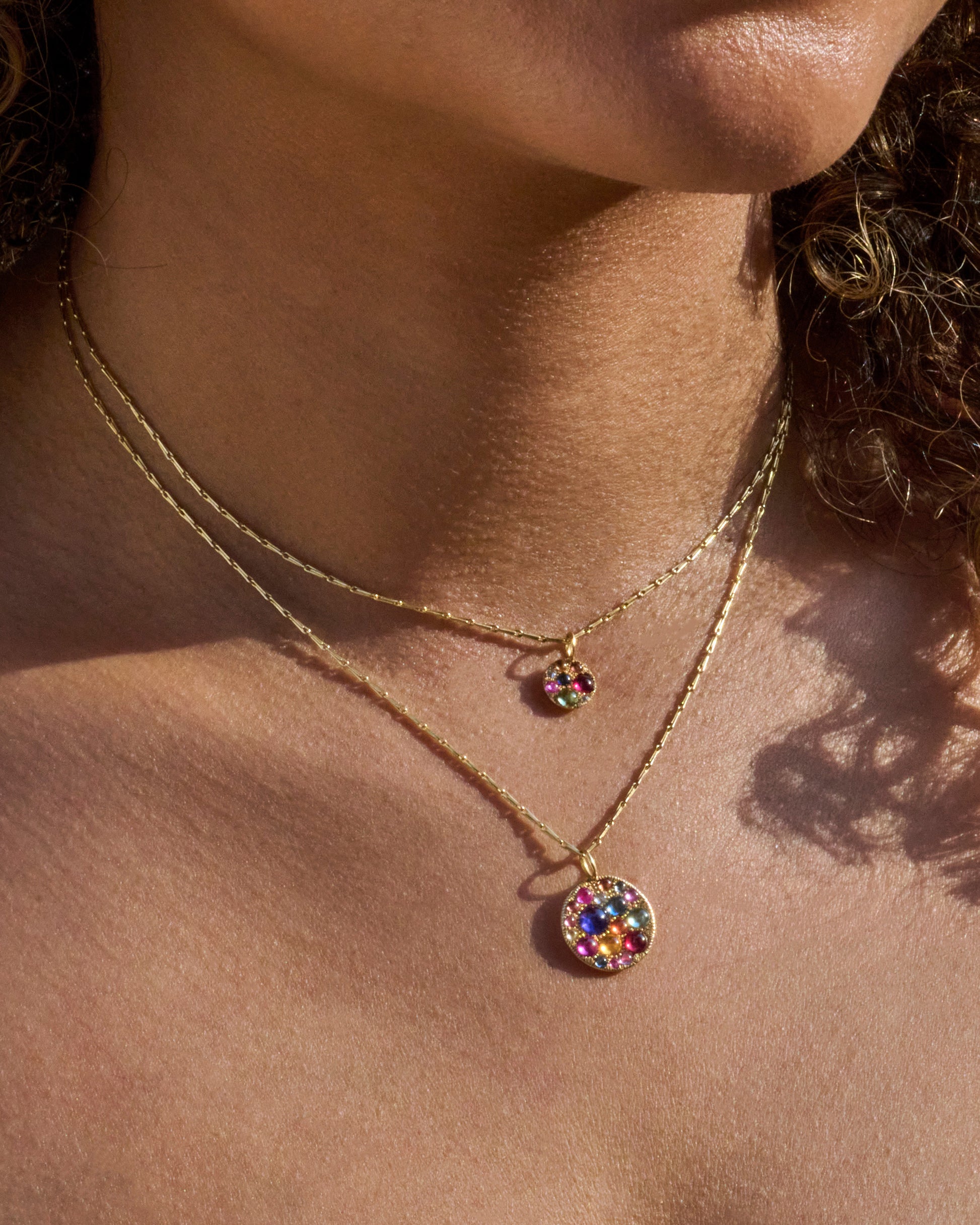 Two yellow gold necklaces, each with a multicolored sapphire and diamond pendant, shown from the front on a person.