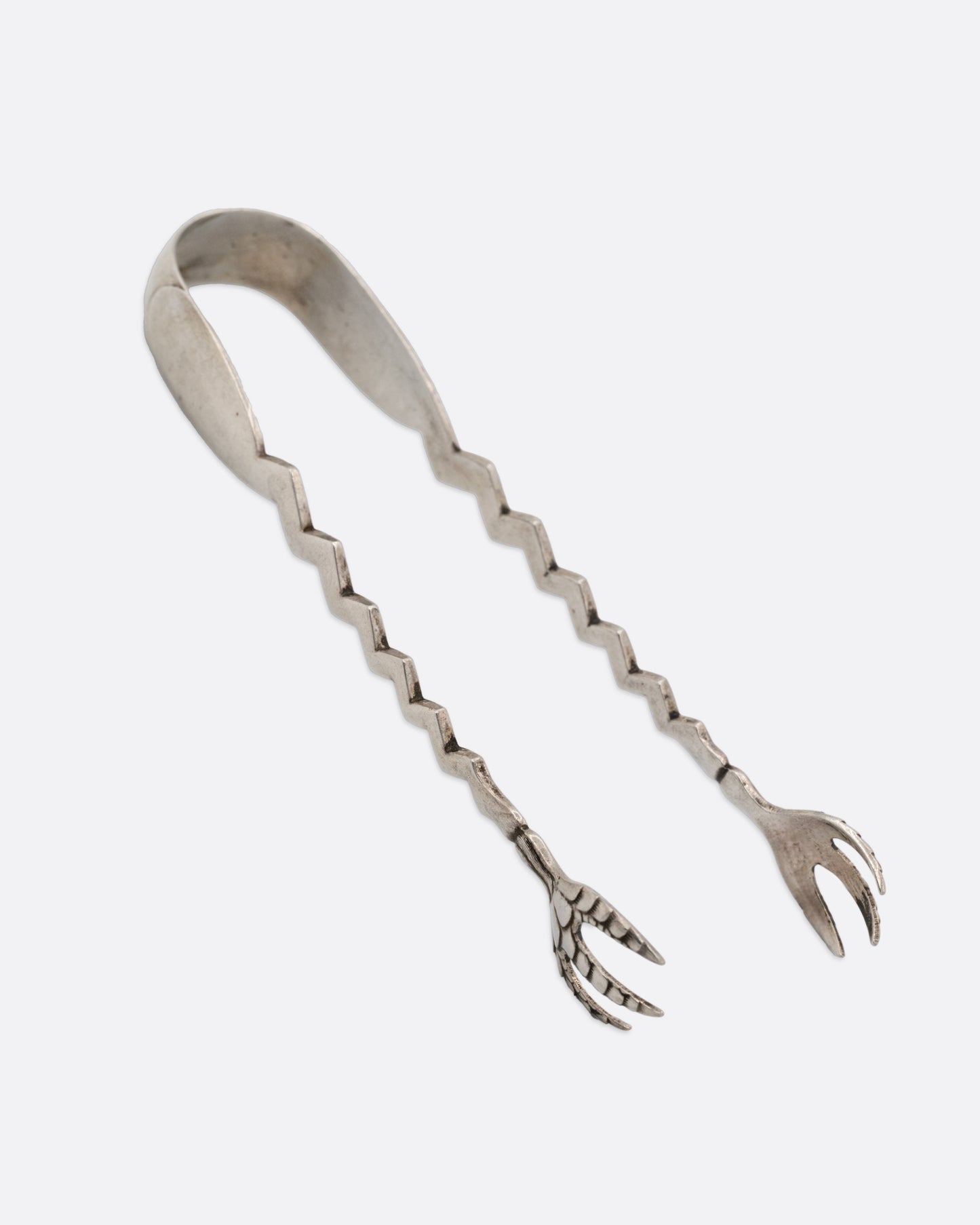 A pair of silver tongs with an arched top, zigzag arms, and claw like 'hands', shown from the side.