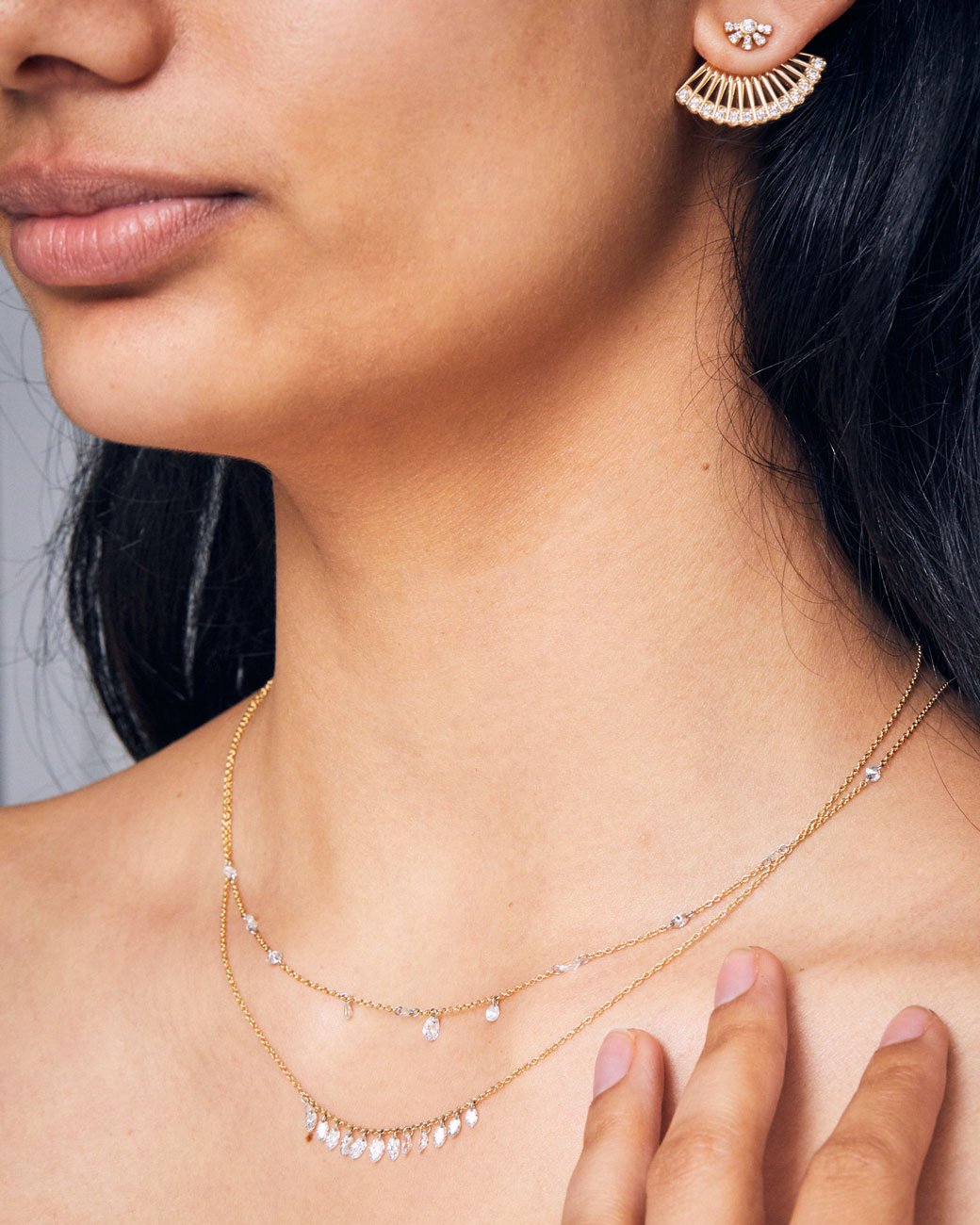 A yellow gold ear jacket shaped like a fan, with diamonds along the bottom edge. Shown from the front on a sun shaped diamond stud earring on a woman's ear. She is also wearing two gold necklaces with diamonds.