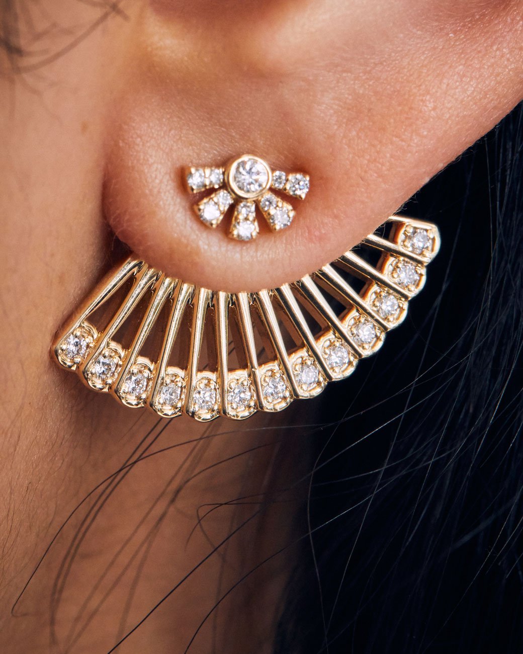 A yellow gold ear jacket shaped like a fan, with diamonds along the bottom edge. Shown from the front on a sun shaped diamond stud earring on a person's ear. 