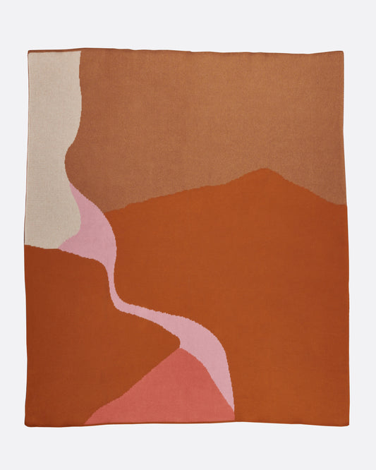 A knit jacquard throw blanket with a tan, brown, and pink motif inspired by the Mojave desert, shown from above.