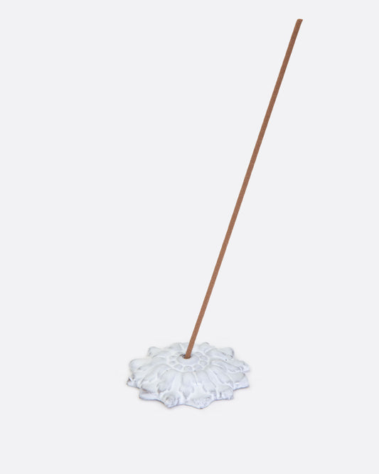 A white incense holder in the shape of a flower with a stick of incense in its center hole. Shown lying flat from the front.