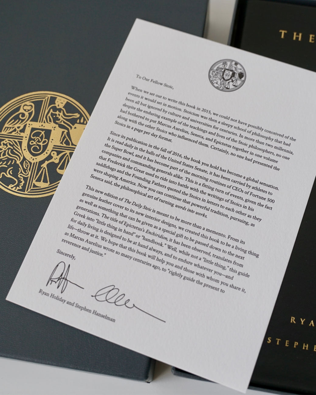 A close up of a letter from the authors of Daily Stoic book, resting atop the book and the box it comes in. The page is white with black lettering. Shown at an angle from the front.