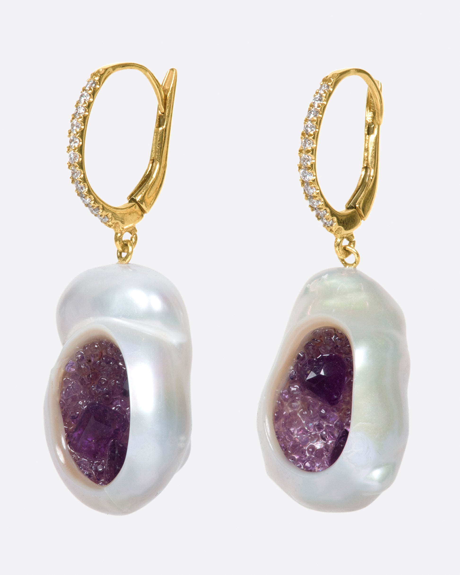A pair of dangle earrings with carved out pearls lined with amethysts. Each earring is topped with  yellow gold ear hook with pave diamonds. Shown from the side.