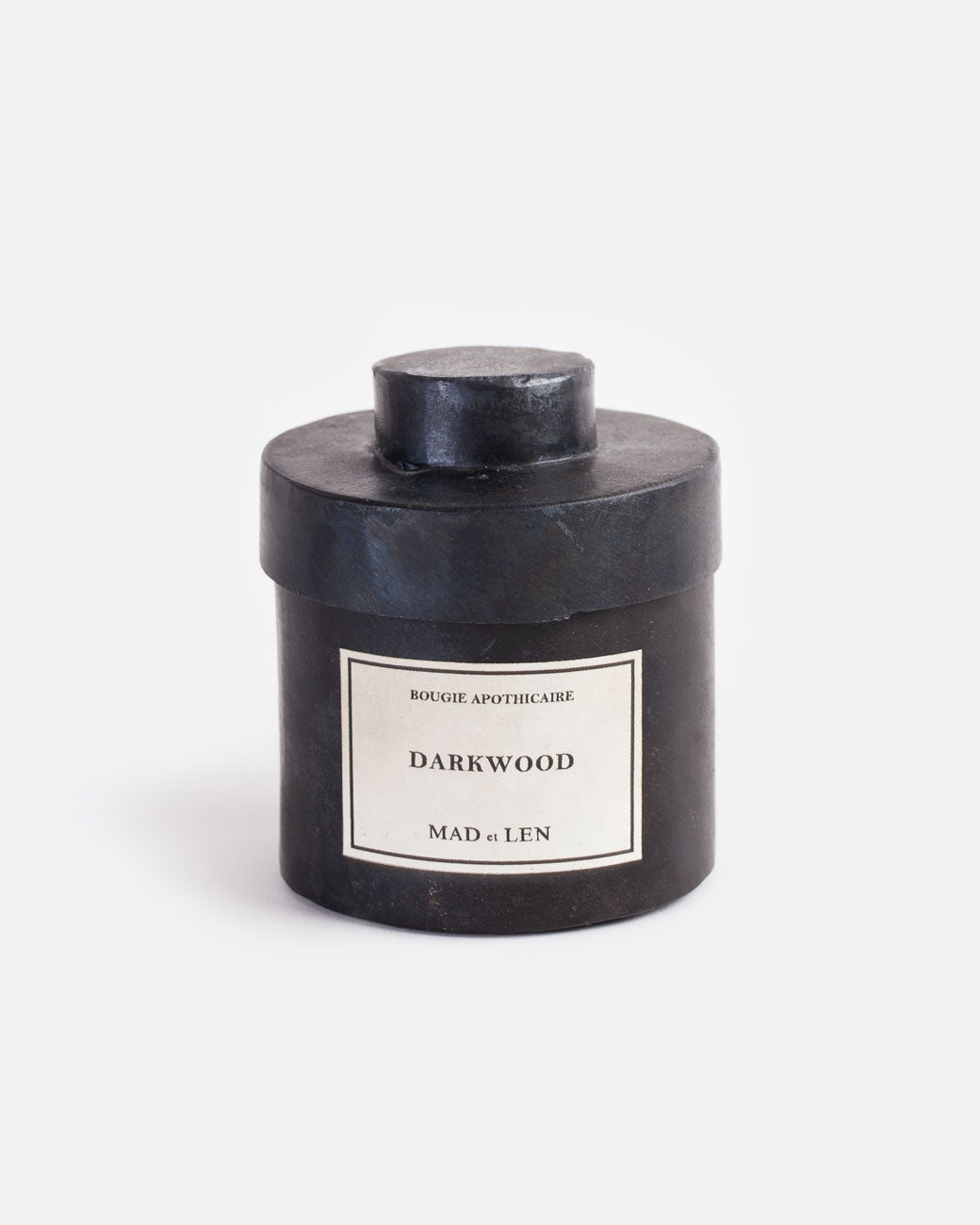A Mad et Len bois debene (darkwood) candle measuring 3" tall and 3.25" wide. It is housed in a one-of-a-kind blackened iron vessel with a lid and a label that reads: Bougie Apothicaire Darkwood Mad et Len. Shown from the front.
