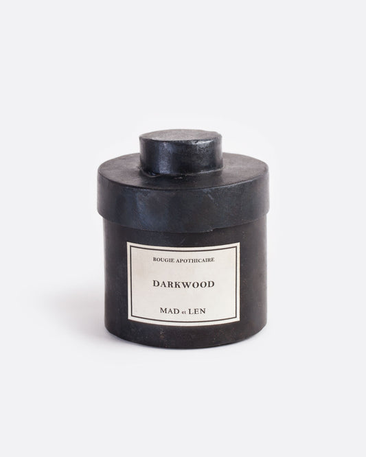 A Mad et Len bois debene (darkwood) candle measuring 3" tall and 3.25" wide. It is housed in a one-of-a-kind blackened iron vessel with a lid and a label that reads: Bougie Apothicaire Darkwood Mad et Len. Shown from the front.