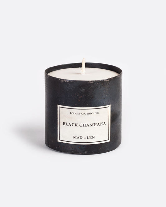 A Mad et Len Black Champaka candle measuring 3" tall and 3.25" wide. It is housed in a blackened iron vessel with a label that reads: Bougie Apothicaire Black Champaka Mad et Len. Shown from the front.