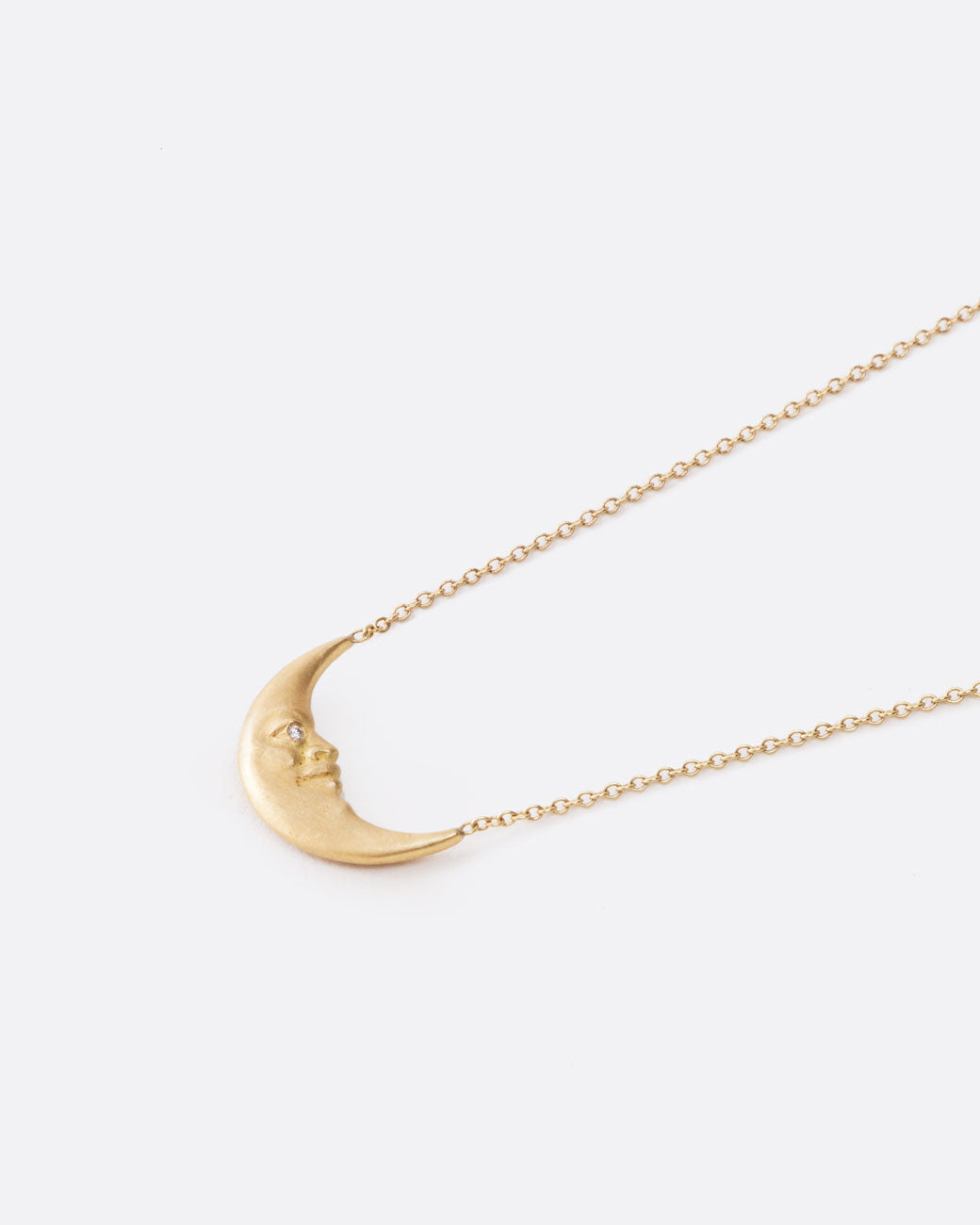 A yellow gold crescent moon pendant necklace with a carved face and diamond eyes. Shown lying flat from the side.