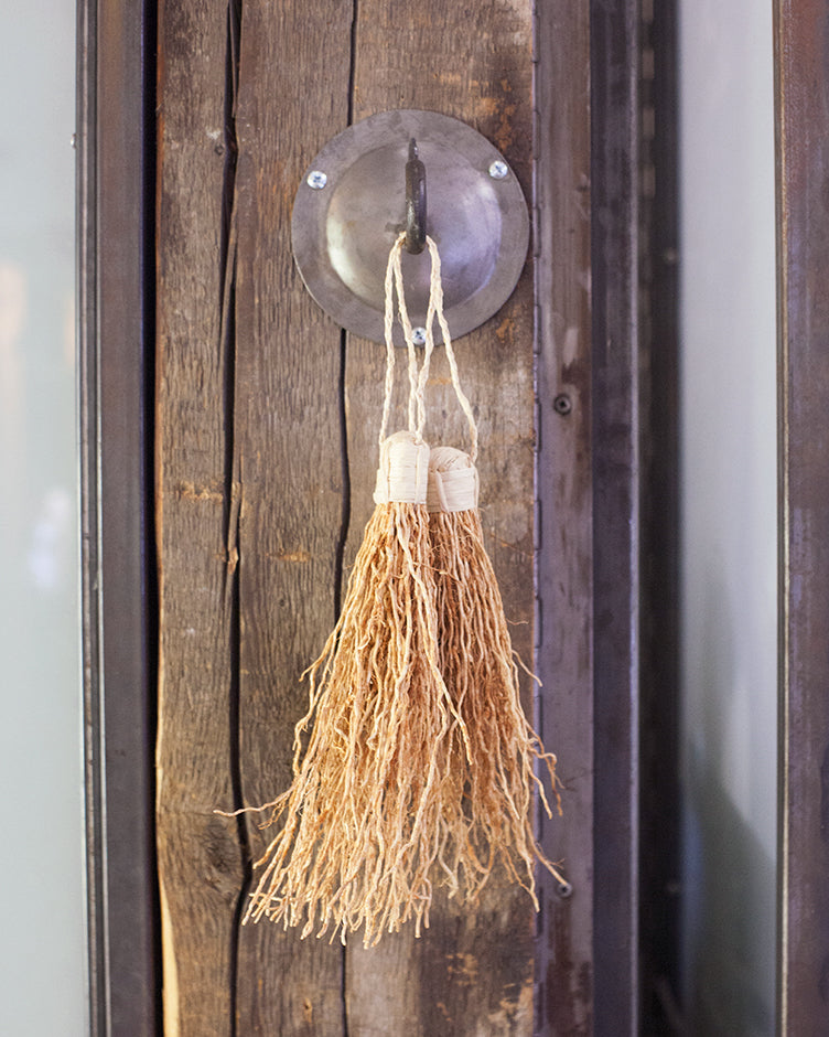 Two vetiver tassels hanging from a metal hook against a wooden beam. Shown from the front.
