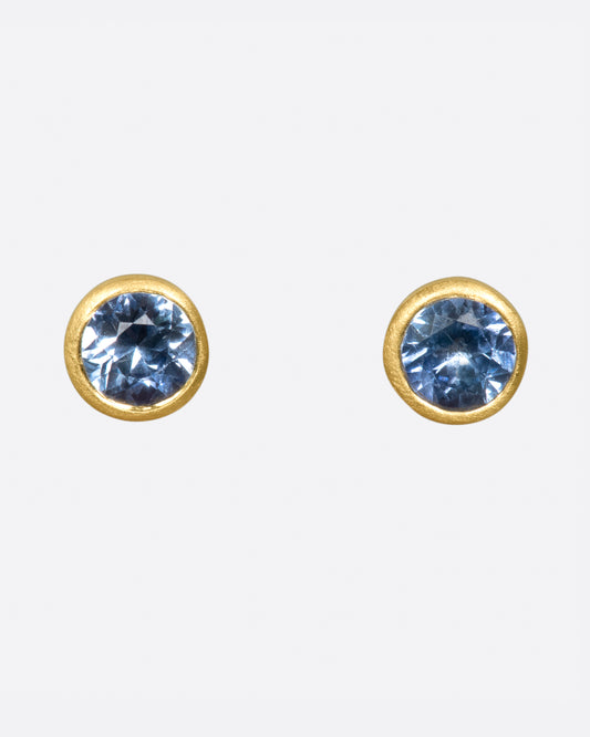 A pair of solitaire earrings featuring medium-blue sapphires in matte gold bezels.
