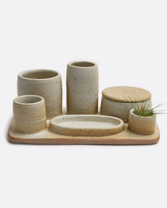 A stoneware pottery organizer tray with various sized cups, jars, and a dish in a matte beige finish. Shown from the front.