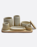 A stoneware pottery organizer tray with various sized cups, jars, and a dish in a matte beige finish. Shown from the front with the jar lid leaning on one side.