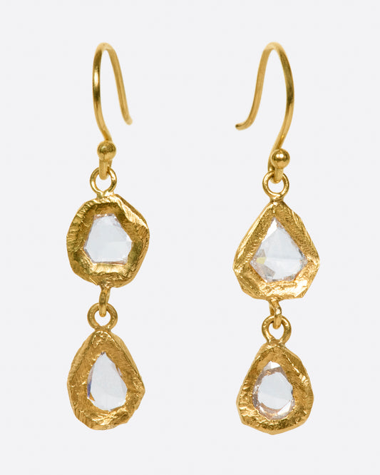 A pair of slightly asymmetrical yellow gold diamond drop earrings with two diamonds set in hand carved bezels on each earring. Shown from the front.