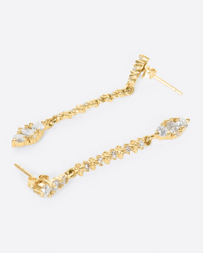A pair of long yellow gold earrings with diamonds throughout. Shown lying on their sides.