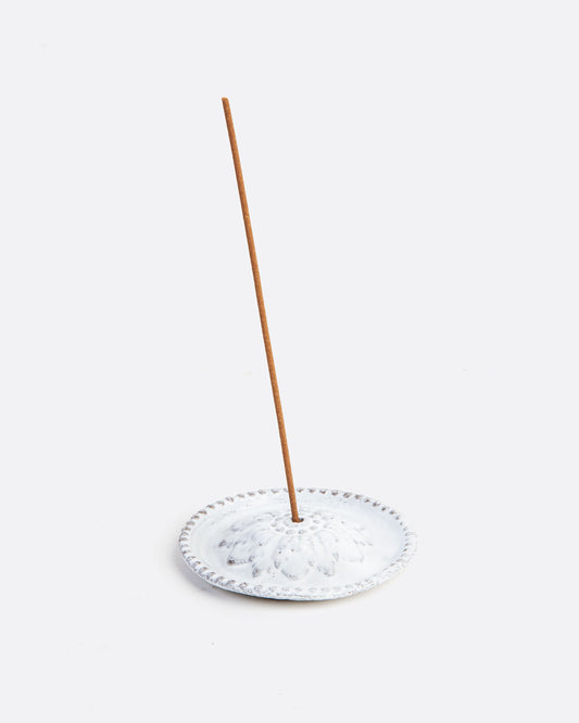 A white ceramic incense holder with a flower at its center and an incense stick in the center hole. Shown from the front.