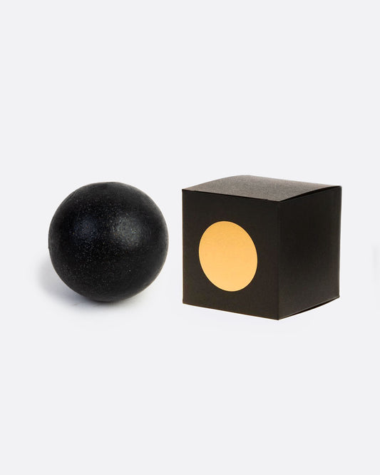 A sphere shaped black soap with the box beside it. The box has a gold circle on the front. Shown from the front.