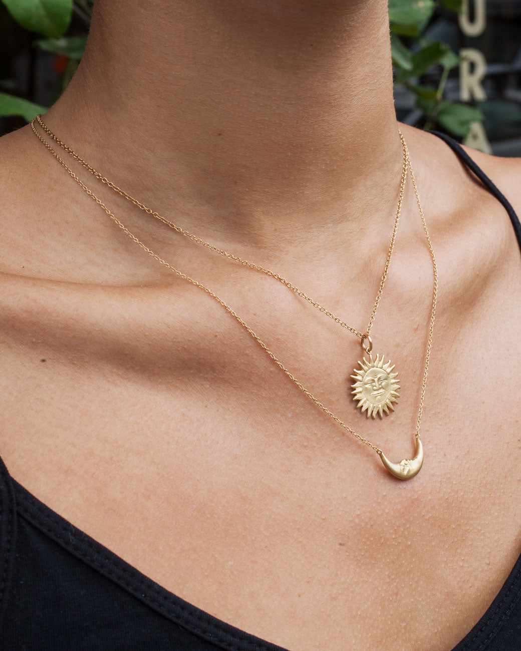 Womans chest wearing two necklaces. The bottom necklace is a yellow gold crescent moon pendant necklace with a carved face and diamond eyes. The top necklace is a yellow gold sun with a carved face. Shown from the front.