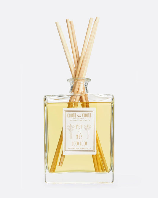 A Coqui Coqui reed diffuser - a glass bottle filled with yellow perfume oil with wooden diffuser sticks out the top and a white label on the front. Shown from the front.